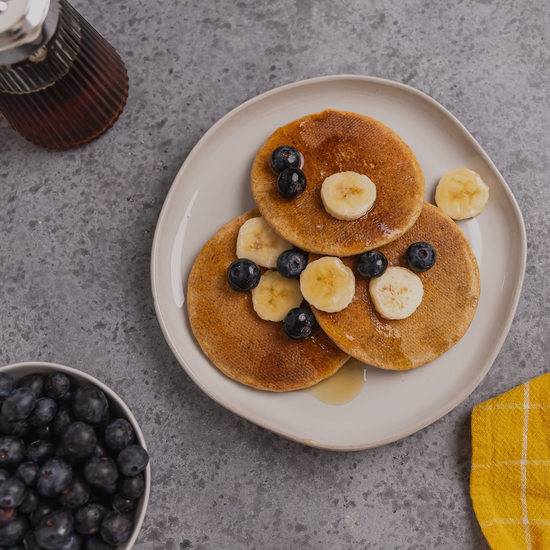 Stack of pancakes with blueberries and bananas on a plate, with syrup and a bowl of blueberries in the background.