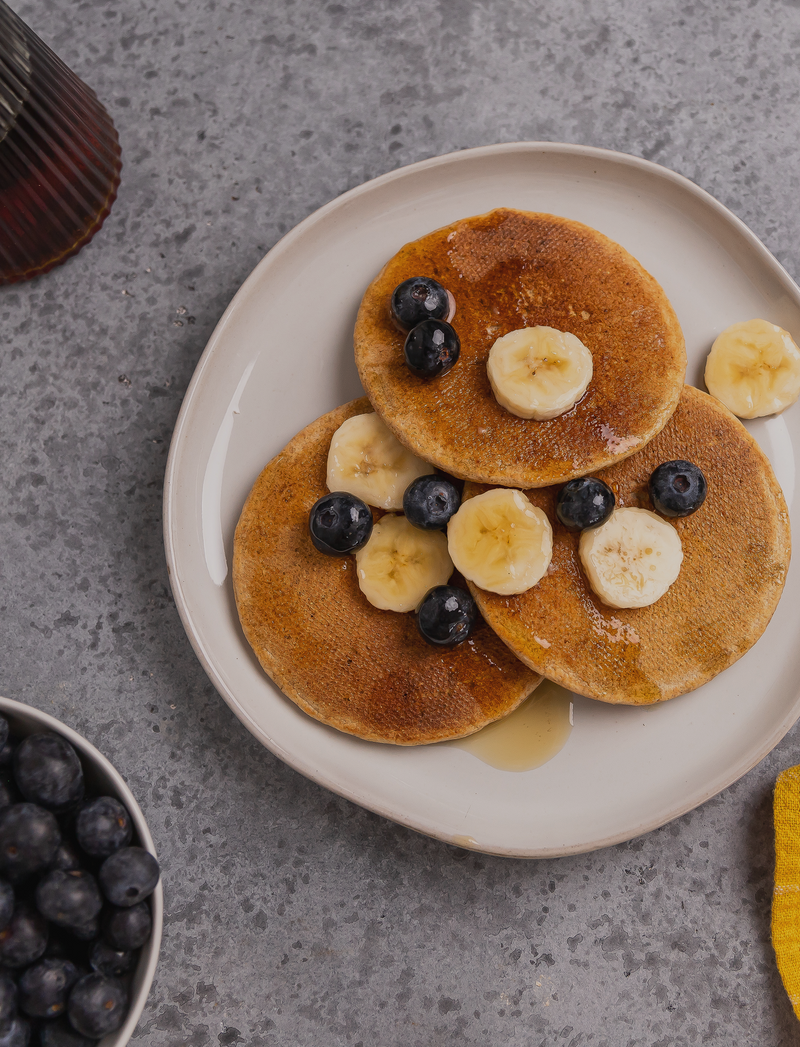 Stack of pancakes with bananas and blueberries on a plate with syrup, on a gray surface.