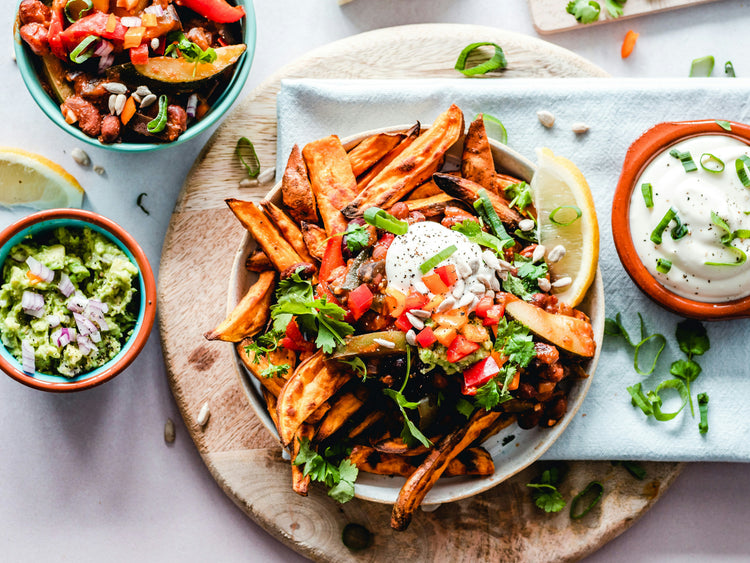 Plated dish of sweet potato fries with toppings, surrounded by small bowls of guacamole, sour cream, and tomatoes on a light background.